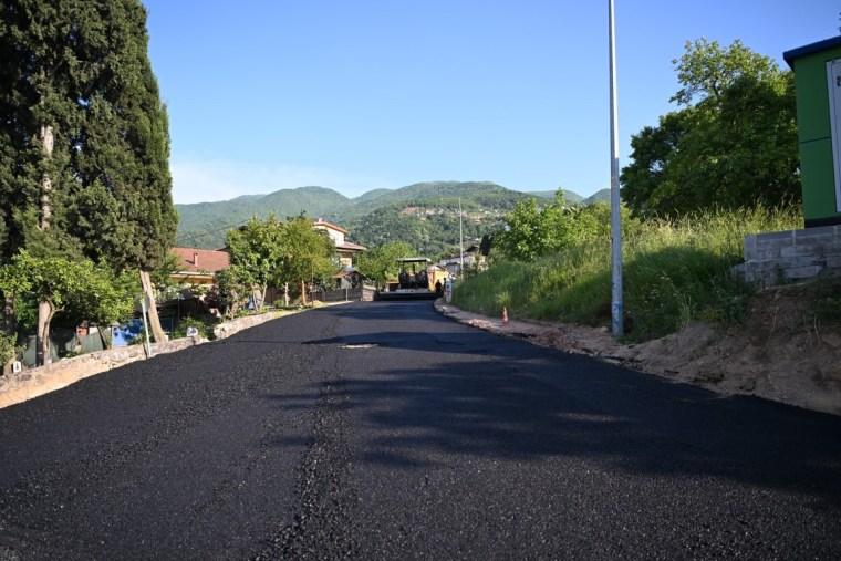 Yeni asfalt serimi yapılan bir yol, yeşil ağaçlar ve dağ manzarası ile çevrili. Güneşli bir gün ve yol yüzeyine sıcak asfalt dökülüyor.