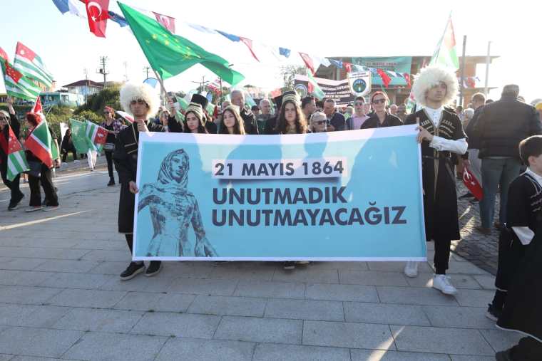 A group of people holding a large banner that reads '21 MAYIS 1864 UNUTMADIK UNUTMAYACAĞIZ' during a public gathering, with flags from various countries in the background.