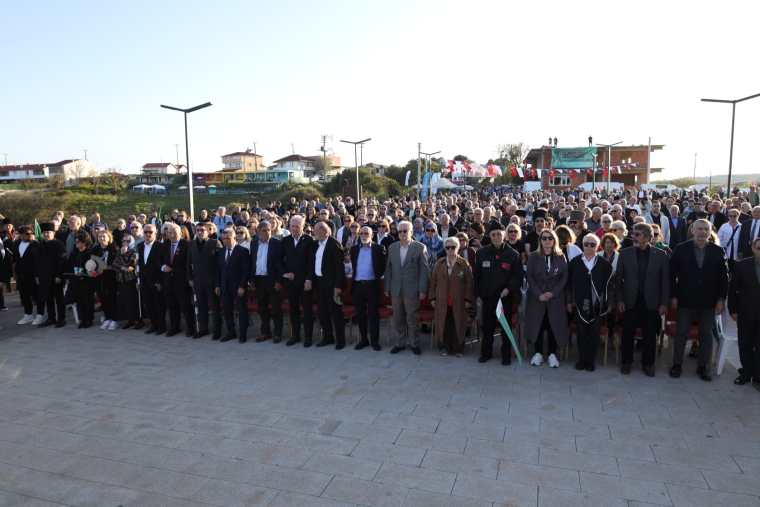 A large crowd of people standing outdoors during a ceremony, with various individuals dressed in formal and casual attire, some wearing glasses. The background includes buildings and flags.