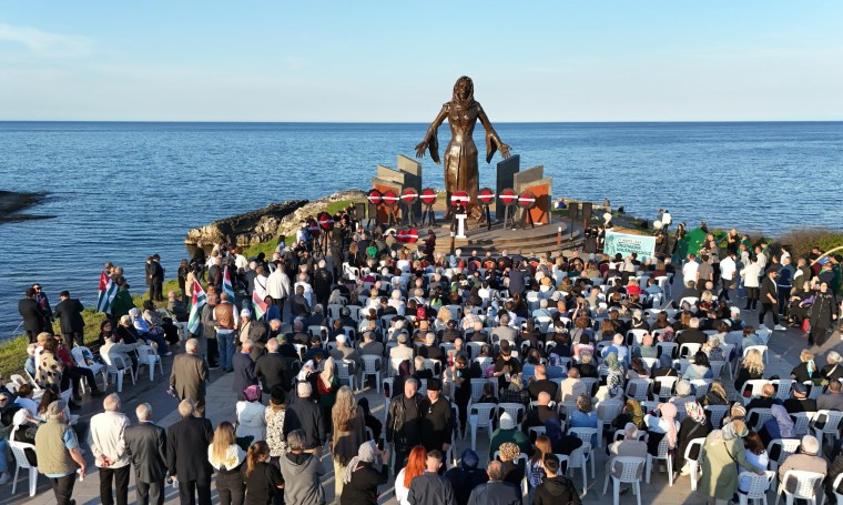 A large crowd gathers near the ocean for a ceremony, with a prominent statue in the background. The audience is seated in white chairs, and various flags are displayed.
