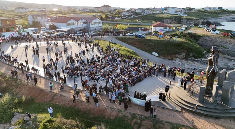 Aerial view of a large crowd gathered at a coastal monument, with houses and greenery in the background and a statue visible in the foreground.