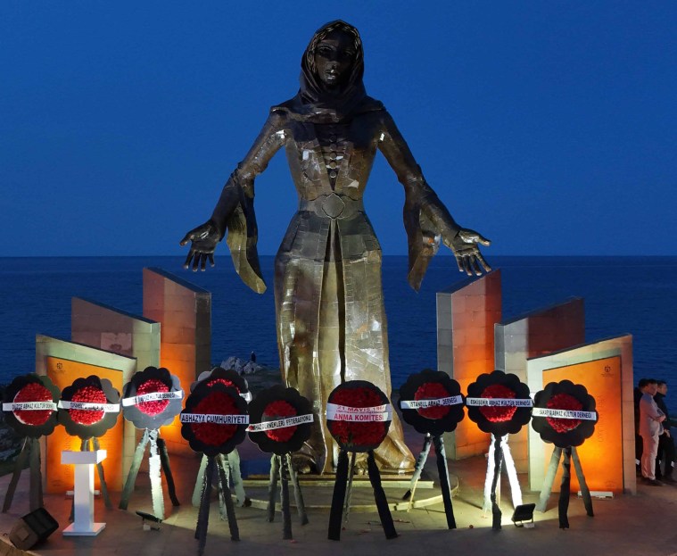 A large metallic statue of a woman with outstretched arms stands by the sea, surrounded by floral tributes on stands at dusk.