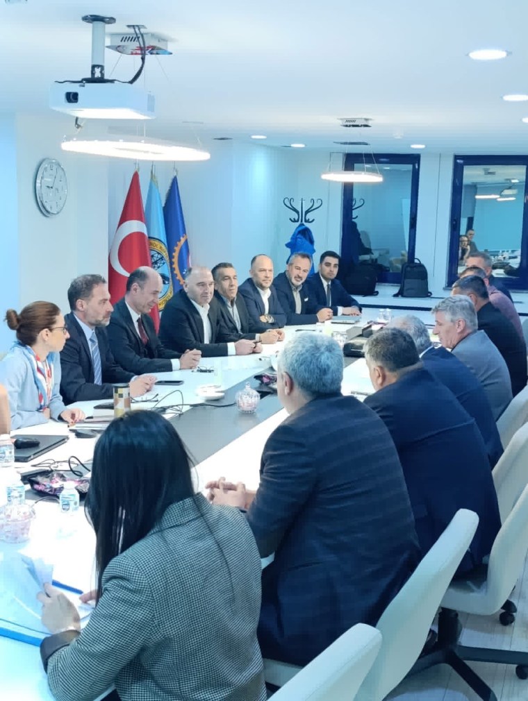 Bir toplantıda oturan insanlar, masanın etrafında işçi sendikası ve yönetim temsilcileriyle müzakerede bulunuyorlar. Arka planda Türk bayrağı ve diğer bayraklar görünmektedir.
