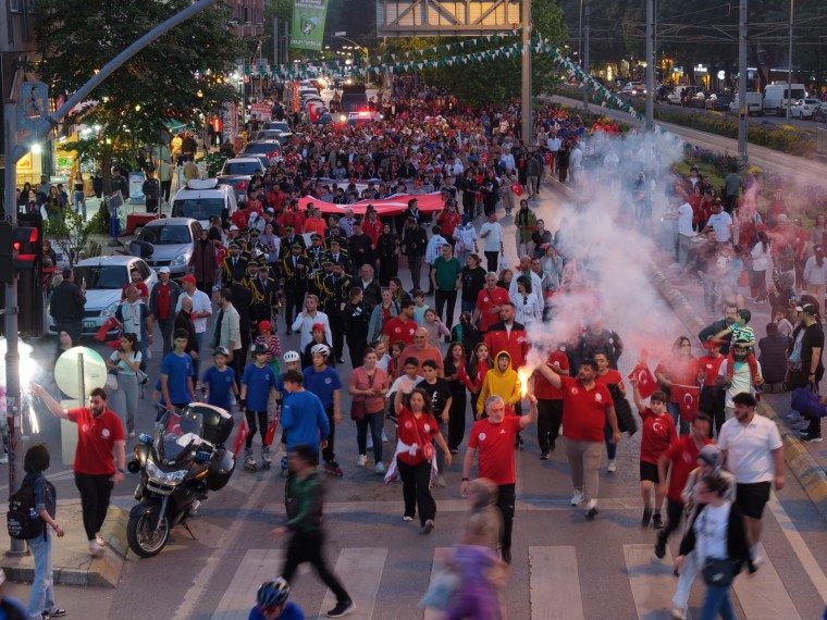 Kalabalık bir grup, sokakta Türk bayraklarıyla 19 Mayıs kutlamaları için yürüyüş yapıyor. Renkli kıyafetler giymiş insanlar, coşkulu bir atmosfer oluşturuyor.