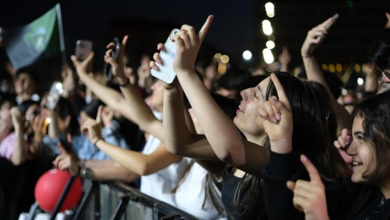 Kalabalık bir gençlik, elleriyle havaya kaldırılmış telefonlar ve işaret parmağıyla 1 işareti yaparak coşkulu bir konser atmosferi yaşıyor.