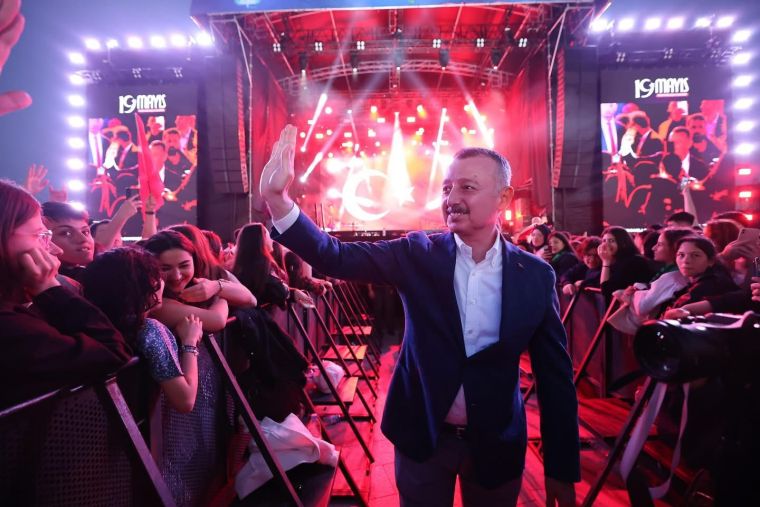 Bir etkinlik sırasında kalabalığa el sallayan bir adam, arka planda sahnedeki ışık gösterileriyle. Etkinlik, 19 Mayıs coşkusunu kutlamak amacıyla düzenlenmiş.