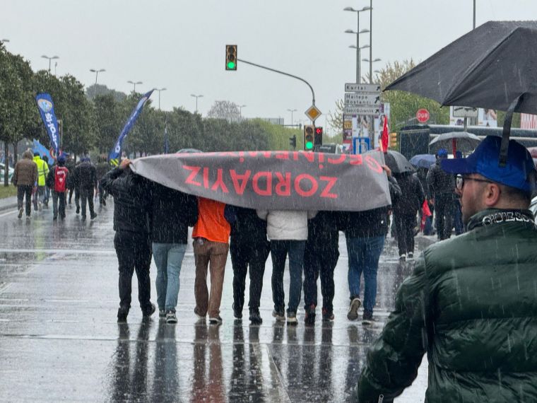 Yağmurlu bir günde, TÜPRAŞ işçileri, 'Zordayız' yazılı büyük bir pankartı taşırken yürüyüş yapıyor. Arkada başka katılımcılar ve yön levhaları görünmekte.