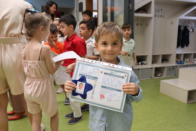 Bir çocuk, karne töreninde elinde karnesiyle gülümseyerek poz veriyor. Arkada diğer çocuklar karne alıyor.