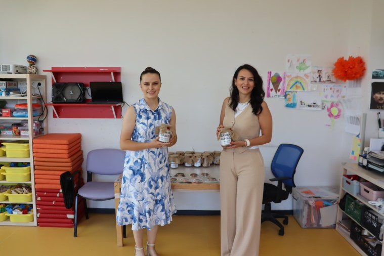 İki kadın, Çınar Çocuk Evleri'nde düzenlenen karne töreninde zaman kapsülü tutarak gülümsüyor. Arka planda eğitim materyalleriyle dolu bir raf ve çeşitli oyuncaklar yer alıyor.