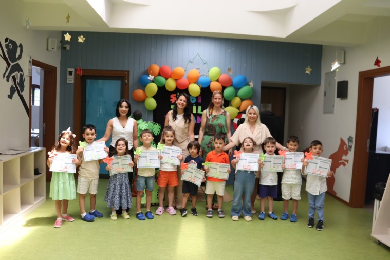 Çınar Çocuk Evleri'nde düzenlenen karne töreninde, öğretmenler ve çocuklar birlikte fotoğraf çektiriyor. Arka planda renkli balonlarla süslenmiş bir alan görünmekte.