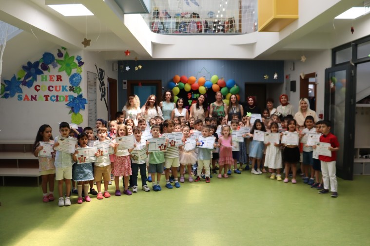 Bir grup çocuk, Çınar Çocuk Evleri'nde karne töreninde yer alıyor, karnelerini gururla tutarken gülümsemekte. Arka planda renkli balonlarla süslenmiş bir duvar görülebiliyor.