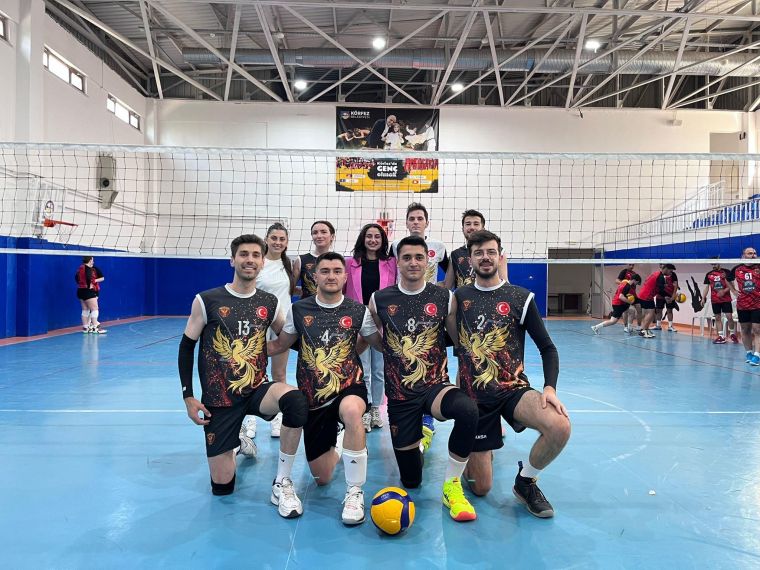 Voleybol turnuvasında yer alan bir takım oyuncularının poz verdiği sahne. Arka planda diğer oyuncular antrenman yapıyor.