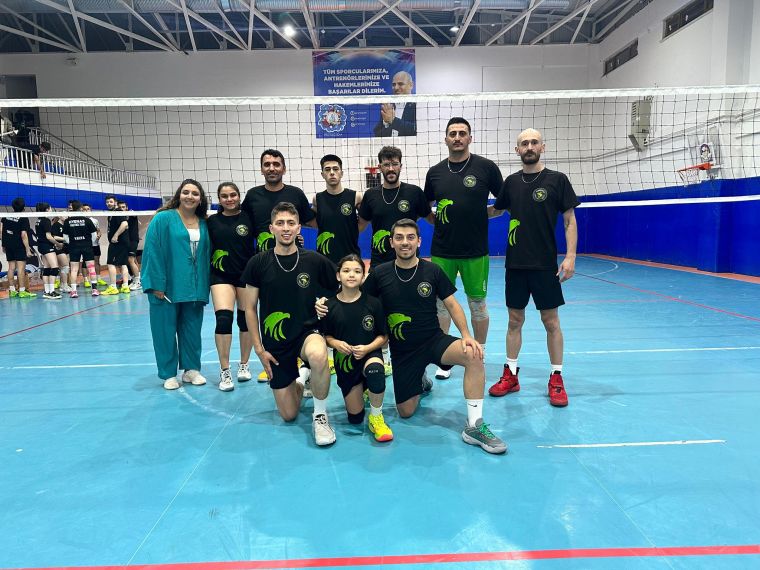 Bir voleybol takımının, turnuvada gülümseyerek poz verdiği bir grup fotoğrafı. Takım üyeleri siyah tişörtler giyiyor ve iç mekanda bir voleybol sahasında duruyor.