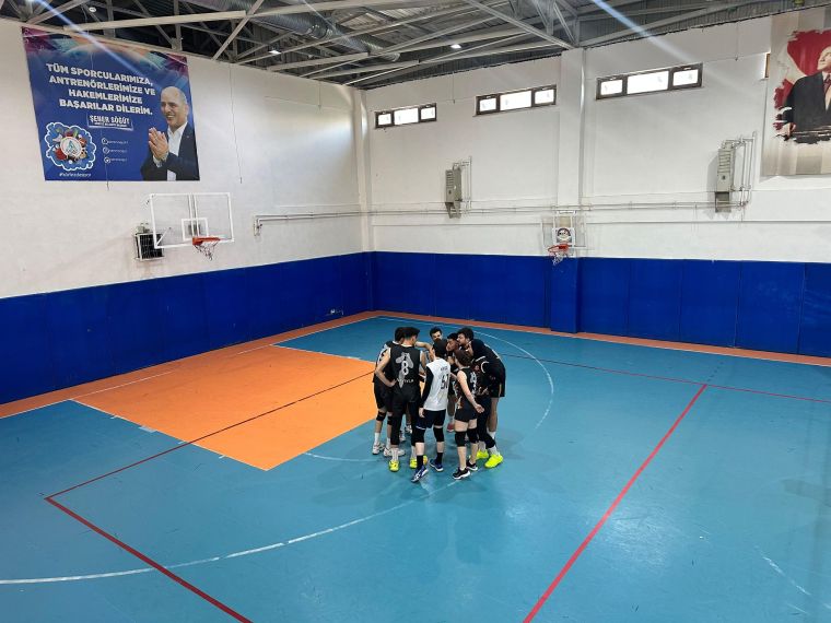 Voleybol takımı, bir spor salonunda maça hazırlanan oyunculardan oluşuyor, takım üyeleri strateji tartışırken halkanın etrafında toplanmış durumda.