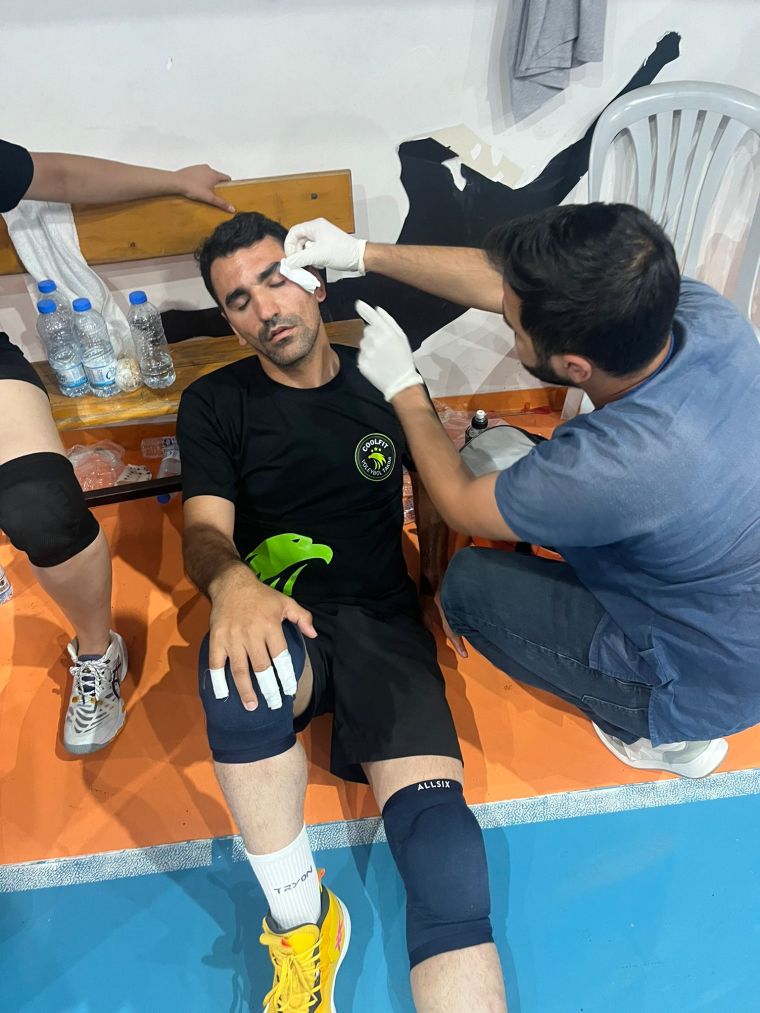 Bir voleybol oyuncusu, yaralanmış yüzüyle bench'te otururken tedavi ediliyor, arka planda su şişeleri var.