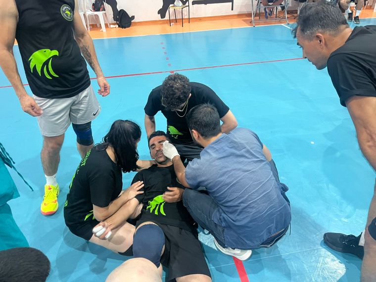 Bir voleybol maçında, yerde yatan bir oyuncuya üç kişi ilk yardım yapıyor. Sporcu, başına bir bandaj sarılıyor ve etrafında destek için birkaç takım arkadaşı bulunuyor.