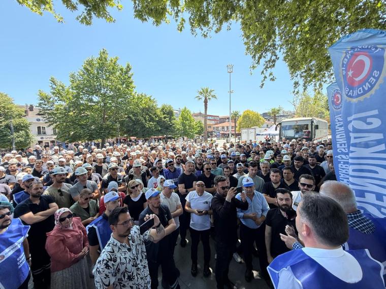 Bir grup işçi, Gölcük Anıtpark'ta toplanmış, eylem yaparak maaş adaletinin sağlanması ve destek çağrısında bulunuyor. Arka planda ağaçlar ve palmiyeler var.