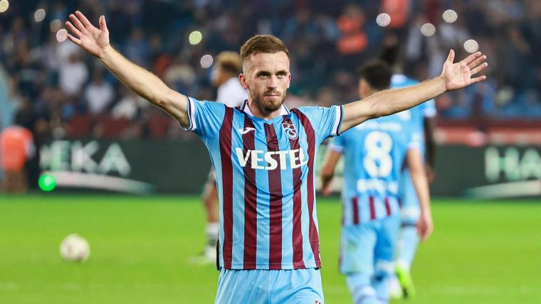 Trabzonspor forması giyen bir futbolcu, stadyumda kollarını açarak taraftarlara selam duruyor.
