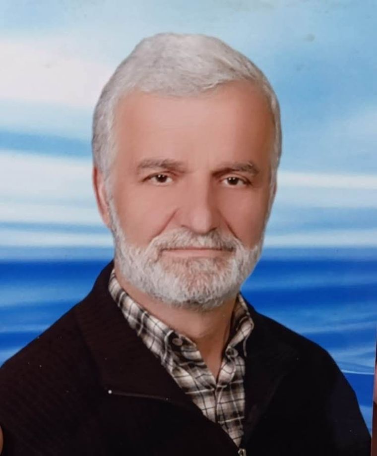 Bir erkeğin, deniz manzarası arka planında gülümseyen portre fotoğrafı. Beyaz saçlı ve sakallı, karalı bir ceket giymektedir.