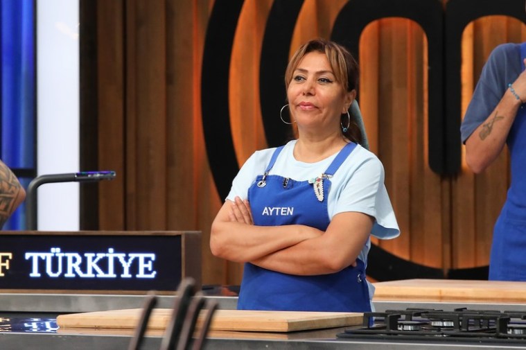 Ayten Saner, MasterChef Türkiye 2025-2026 sezonunda yarışmanın stüdyosunda duruyor, mavi önlük giyiyor ve düşünceli bir şekilde kollarını kavuşturmuş.