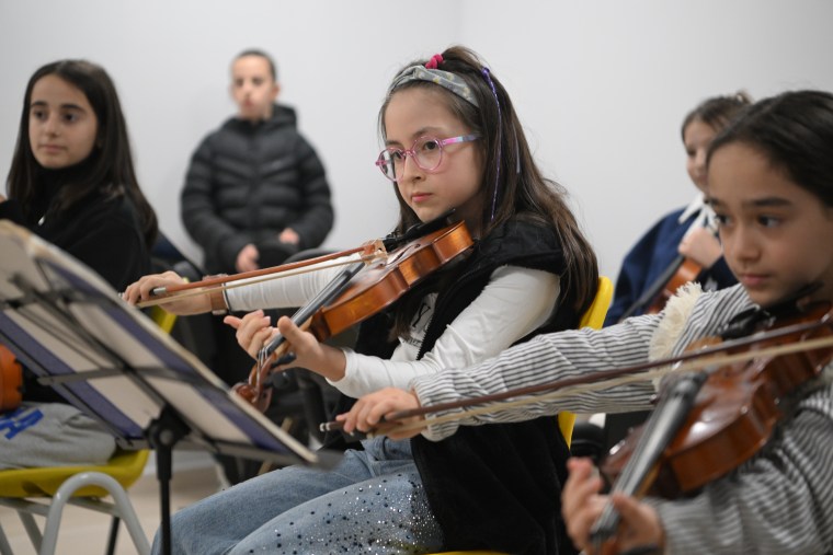 Çocuklar, Kartepe Genç Akademi'de müzik dersi alırken bir arada oturuyorlar. Ön planda bir çocuk, keman çalmakta ve dikkatle dinlemekte.