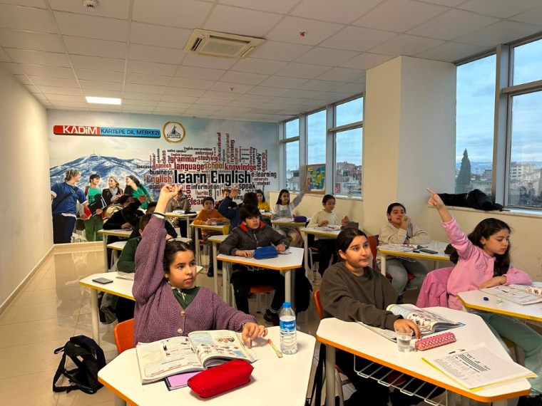 Öğrencilerin İngilizce dersine katıldığı bir sınıf ortamı, öğrencilerin ellerini kaldırarak öğretmene soru sorduğu an.