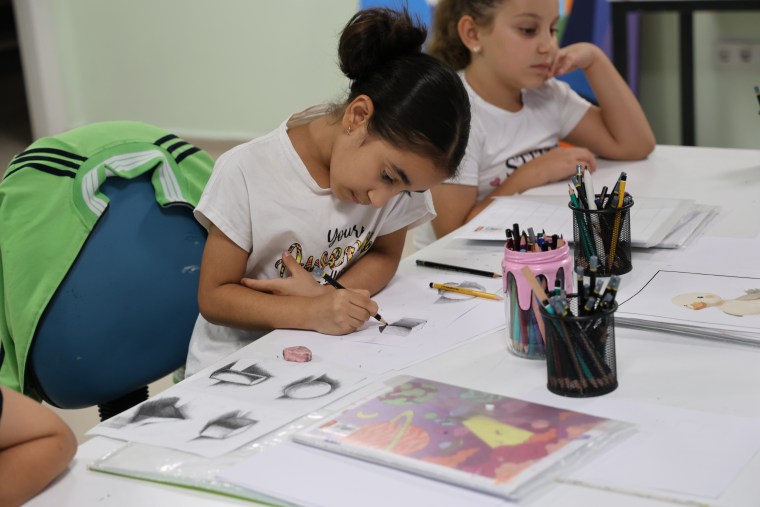 Bir genç kız, sanat atölyesinde çizim yapıyor, masanın üzerinde renkli kalemler ve çeşitli sanat malzemeleri var.