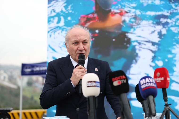 Bir konuşma yapan kişi, arka planda yüzücü görüntüsü olan bir ekranda mikrofon tutuyor.