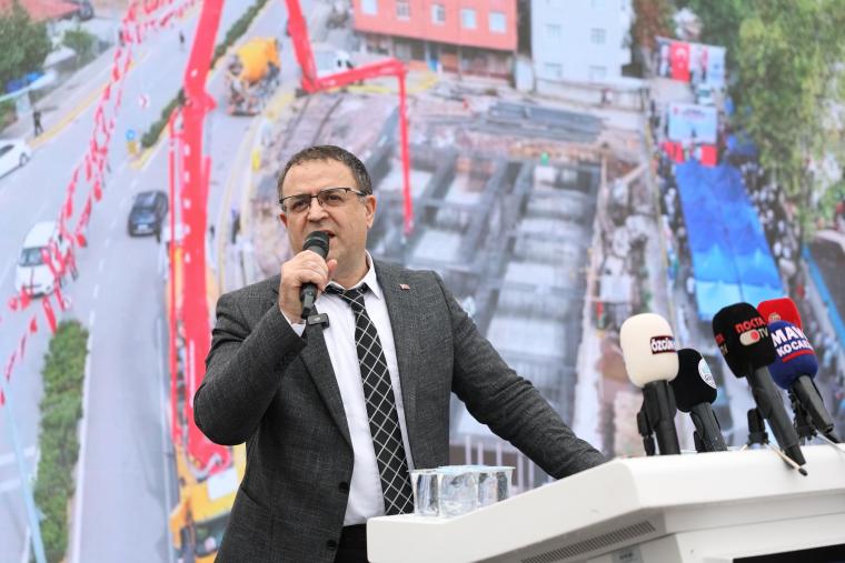 Bir yetkili, arka planda inşaat alanı bulunan bir etkinlikte mikrofonla konuşuyor.