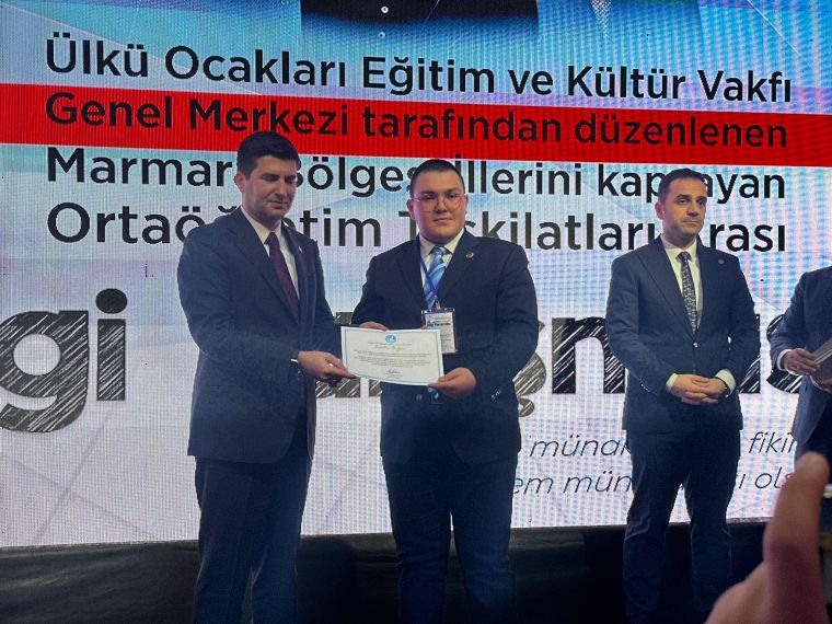 Eğitim ve Kültür Vakfı ödül töreninde sertifika alan bir genç, yanında bir yetkili ile fotoğraf çektiriyor. Arka planda etkinlikle ilgili yazılar yer alıyor.