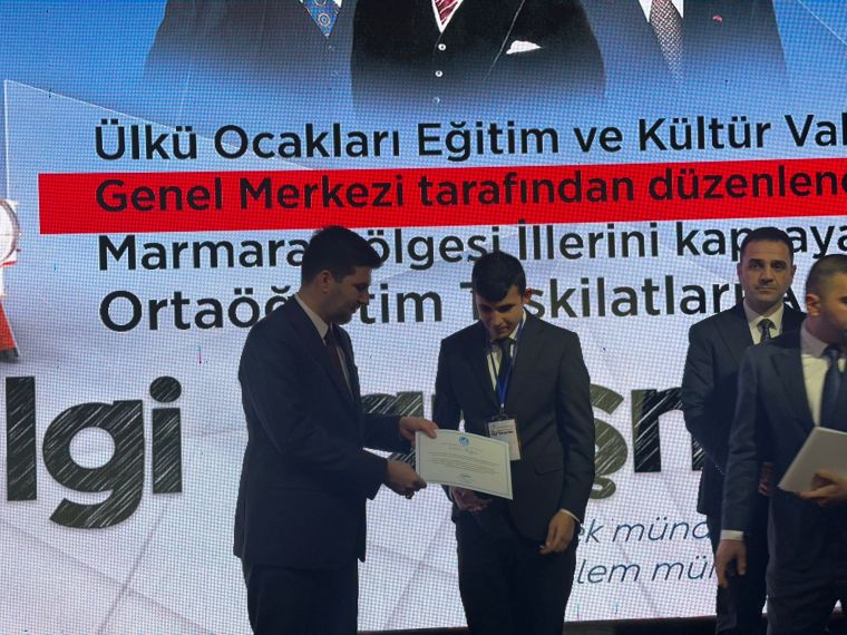 Bir etkinlikte ödül alan bir kişi, belgelerini takdim eden bir yetkili ile el sıkışıyor. Arka planda etkinlik bilgilerini içeren bir ekran bulunuyor.