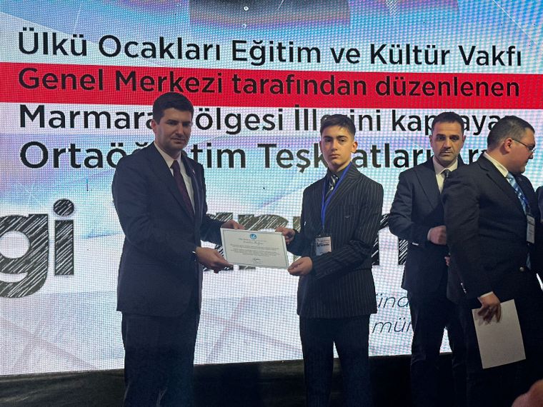 Ödül töreninde bir kişi, sertifika alırken diğerleri izliyor. Arka planda etkinlik detayları yer alıyor.