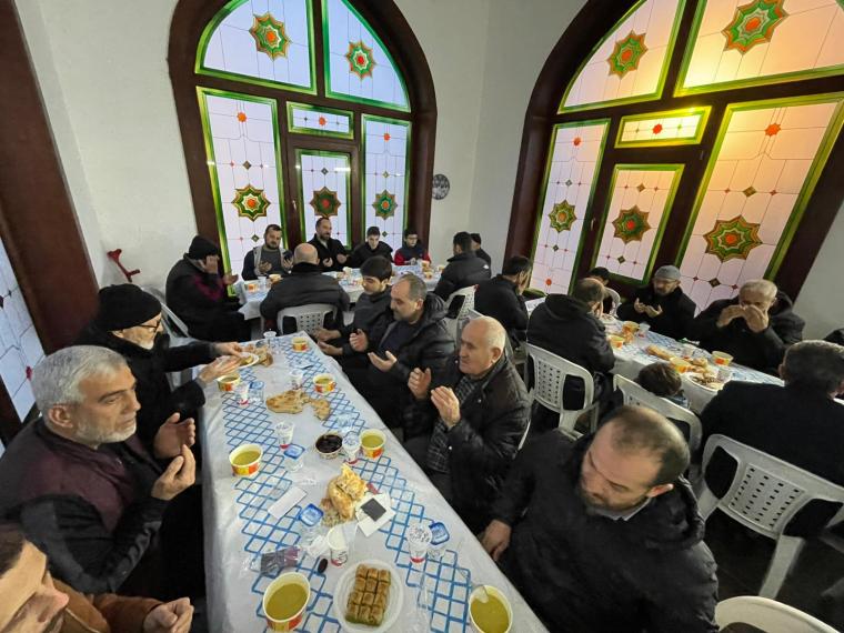 Bir grup insan, bir masanın etrafında oturmuş, dua etmekte ve yemek yemektedir. Mekanın duvarlarında büyük ocak başlığı ve geleneksel desenli pencereler bulunmaktadır.