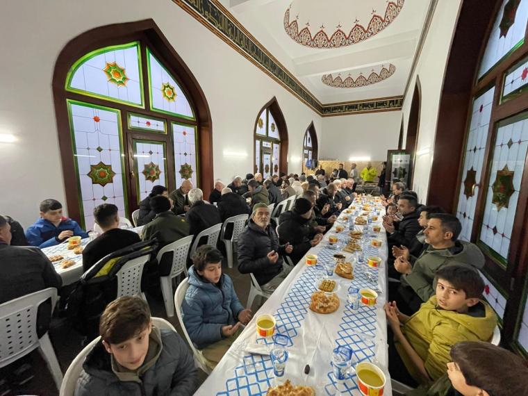 İç mekanda bir yemek masasında oturan kalabalık bir grup insan. Masada yiyecek ve içecekler var, bazıları dua ediyor. Arka planda dekoratif pencereler ve mimari detaylar görünmekte.