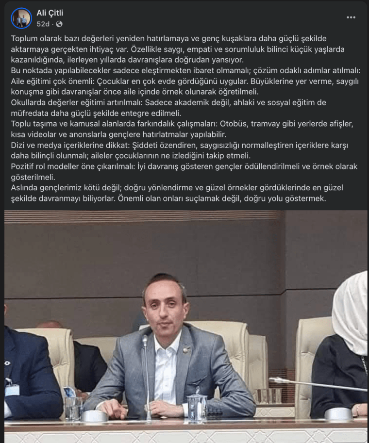 Bir toplantıda konuşan bir adam, masanın arkasında oturuyor. Üzerinde gri bir ceket var ve önünde bir mikrofon ile su bardağı bulunuyor. Arka planda diğer katılımcılar görünüyor.