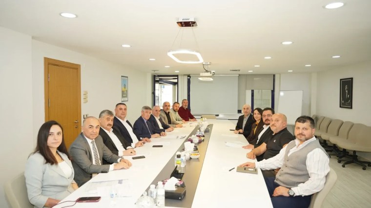 Bir toplantı odasında oturan on dört kişi, uzun bir masa etrafında düzenli bir şekilde sıralanmış. Katılımcılar, resmi bir toplantıya katılıyor gibi görünmektedir.