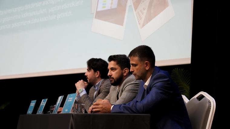 Bir panelde oturan üç konuşmacı, önlerinde mavi belgelerle birlikte sunum yapıyor.